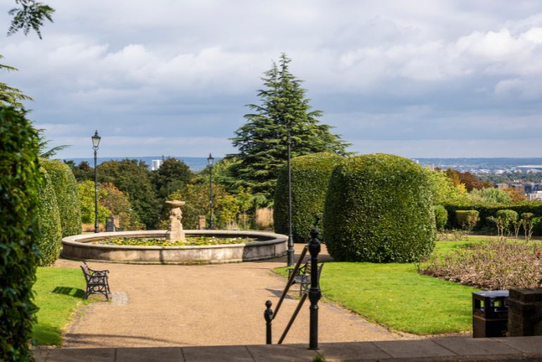 A serene garden with manicured hedges, a central fountain, benches, and a distant view of a sprawling landscape under a partly cloudy sky.