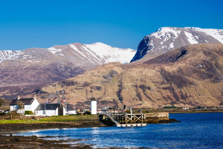 A serene lakeside view with a cluster of white houses, a pier, and snow-dappled mountains under a clear blue sky.