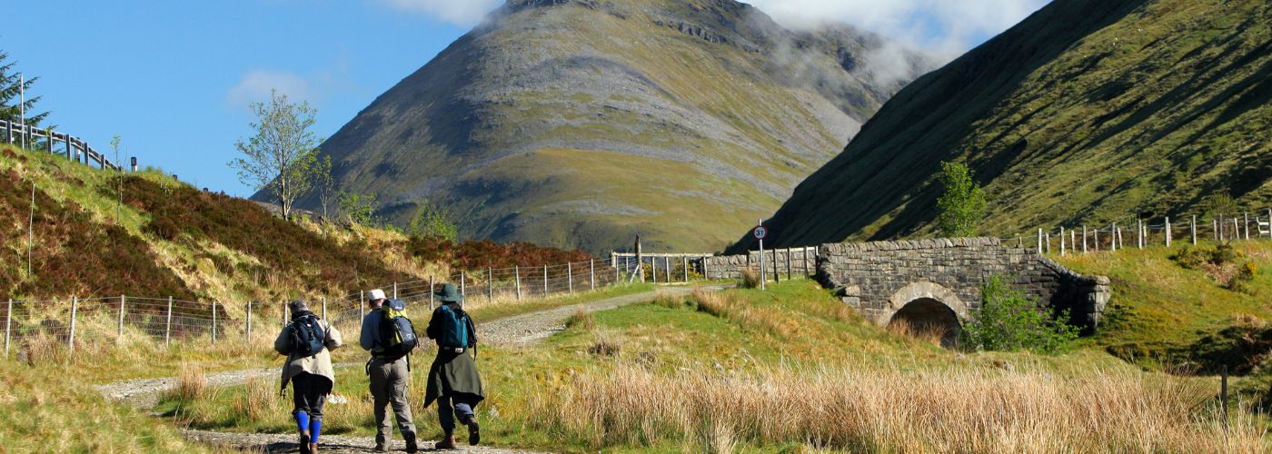 Three hikers with backpacks walking towards a stone bridge in a valley with grassy slopes and a mountain backdrop under a clear sky.