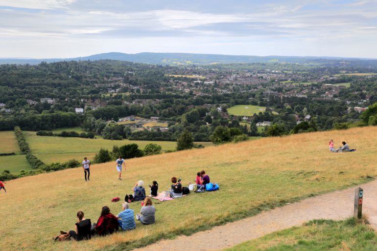 A panoramic view of a lush valley with scattered groups of people sitting on a grassy hillside, overlooking a town amidst greenery under a cloudy sky.