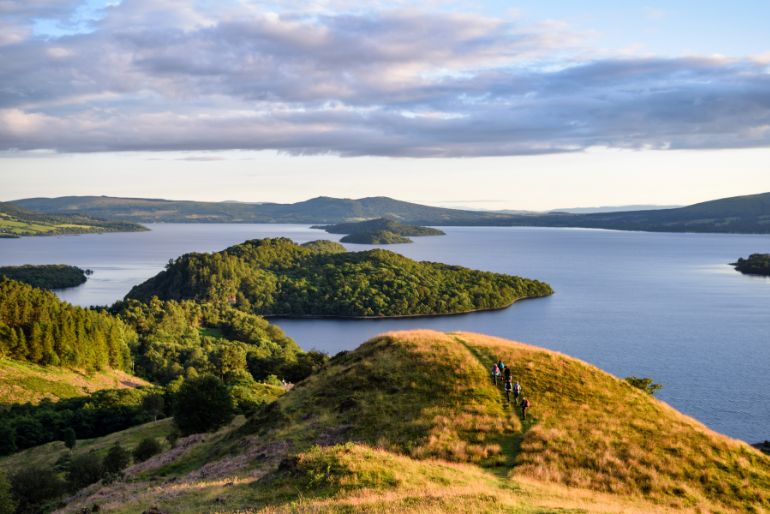 Hills with lush greenery overlook a serene lake with islands under a soft evening light. Hikers are visibly enjoying the tranquil scenery.