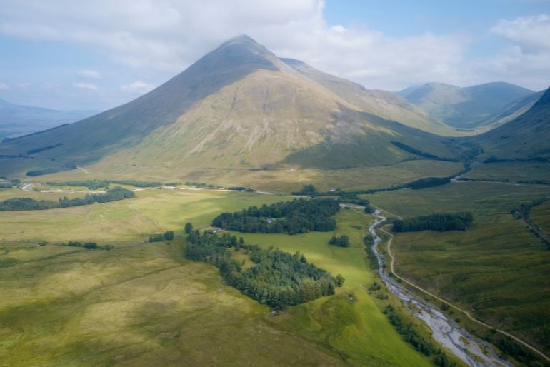 Aerial view of a lush valley with a prominent mountain peak, a winding road, and patches of forest under a partly cloudy sky.