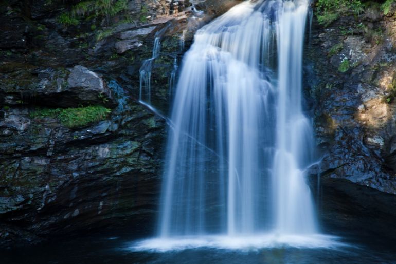 A serene waterfall cascades smoothly over a rocky cliff into a placid pool below, surrounded by lush foliage.