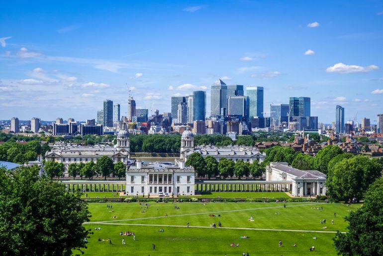 Alt-text: View of a city skyline behind a historical building with a vast green lawn where people are leisurely scattered, under a bright blue sky with fluffy clouds.