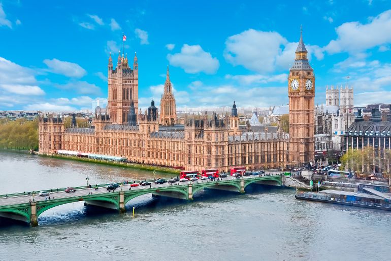 Westminster Palace and Big Ben beside the River Thames with Westminster Bridge, under a partly cloudy sky.