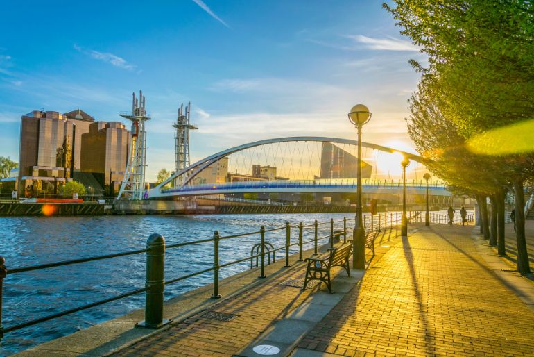 Sunset over a serene waterfront with a curved bridge in the background, lined by trees and a cobblestone pathway in the foreground.