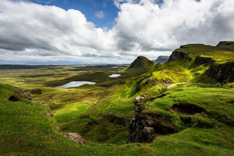 Rolling green hills under a cloudy sky with patches of sunlight, featuring rugged terrain and lakes in the distance.