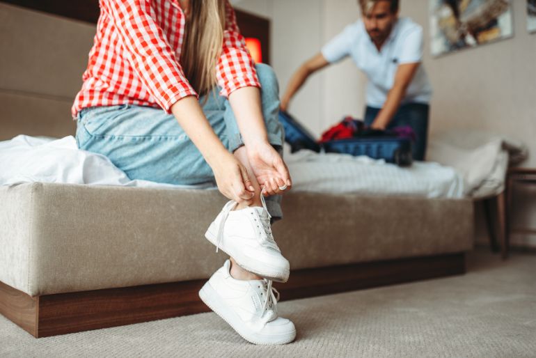 A woman in a red checkered shirt and jeans ties her white sneakers while seated on a bed, with a man packing clothes in the background.