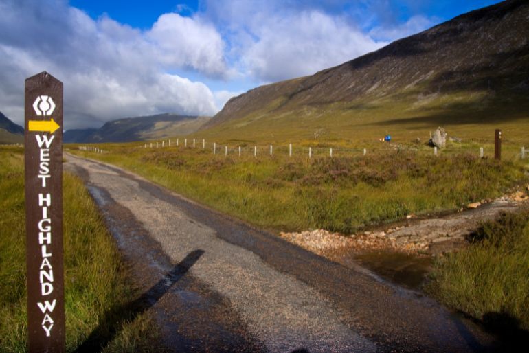 Directional sign for West Highland Way alongside a rural road leading through a serene valley with rolling mountains under a partly cloudy sky.