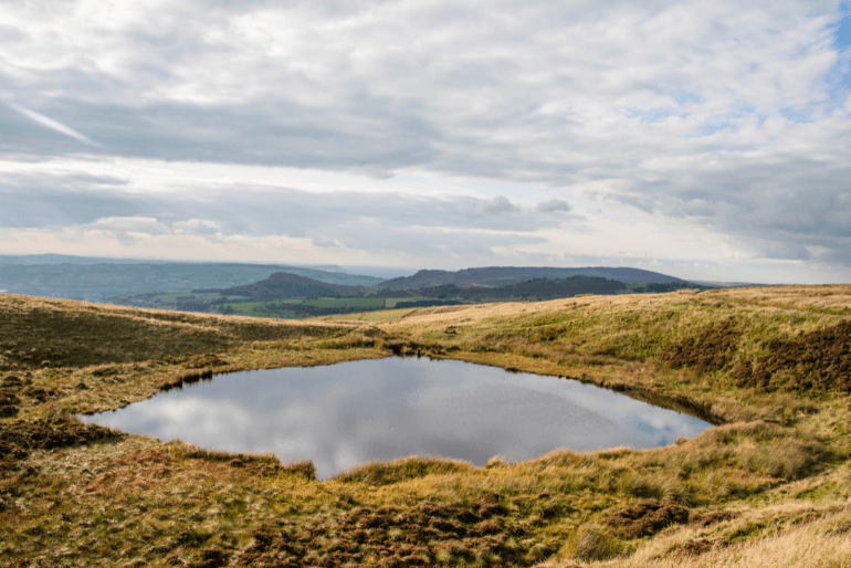 Tranquil expansive valley with a reflective pond surrounded by rolling grassy hills under a partly cloudy sky.