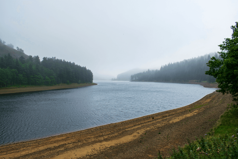 Tranquil lake with stones on the bank, misty sky and lush trees, at upper Derwent valley