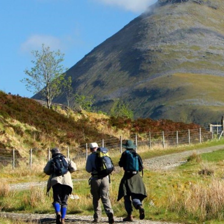 Three hikers with backpacks walking towards a stone bridge in a valley with grassy slopes and a mountain backdrop under a clear sky.