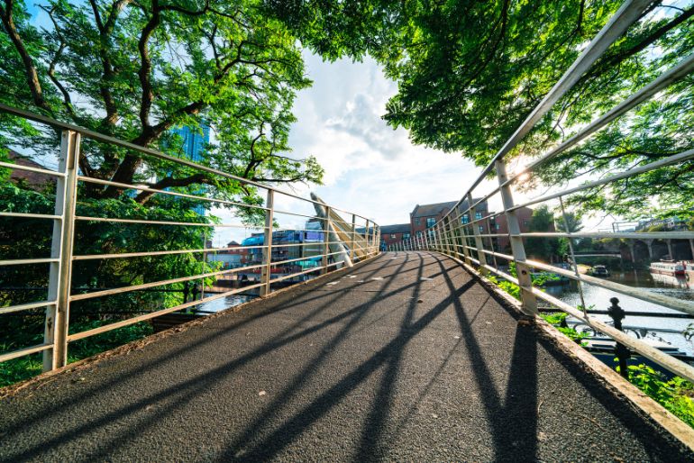 A tranquil footbridge with green trees, casting long shadows over the path, overlooking a calm river and urban structures in the background.