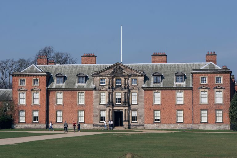 Elegant, red-brick country house with large windows and a central entrance, surrounded by a manicured lawn and visited by people under a clear blue sky.