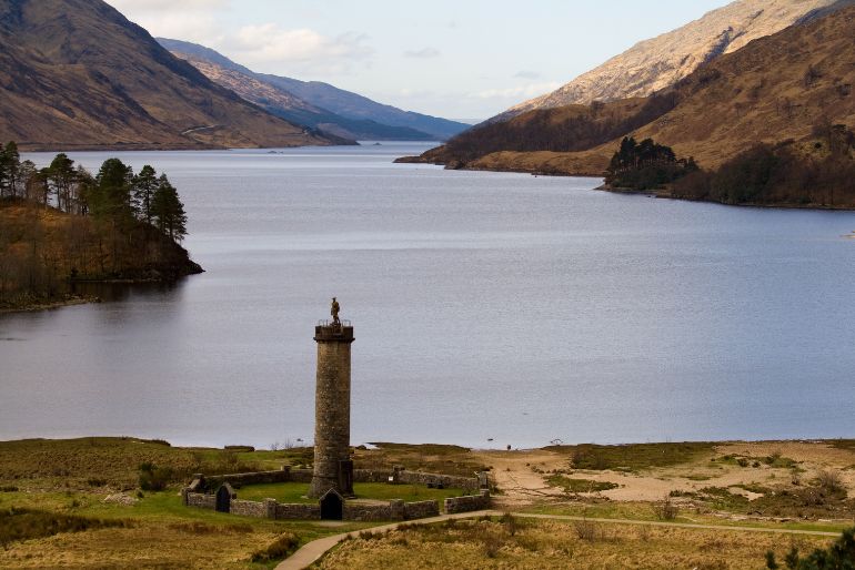 A serene valley with a calm lake, surrounded by rolling hills, featuring a prominent stone monument with a statue atop.