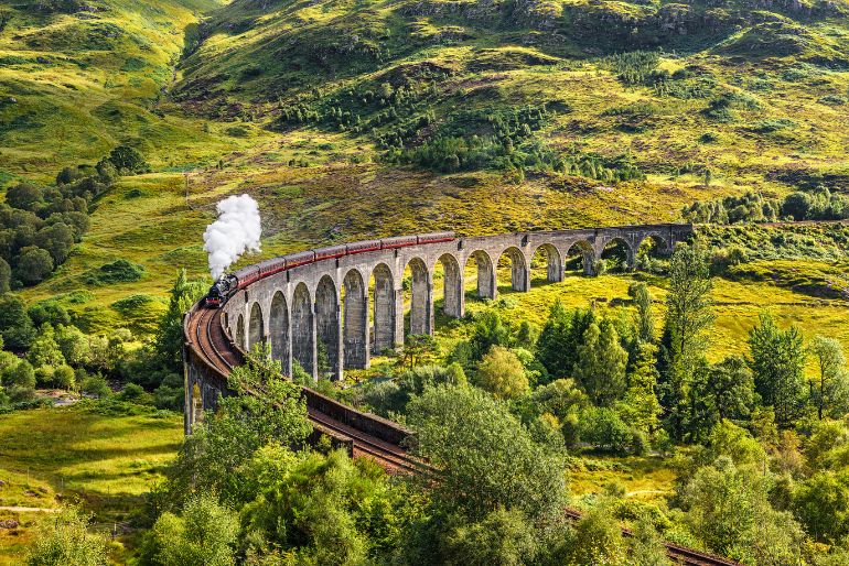 A vintage steam train crosses a long arched viaduct amidst vibrant green rolling hills under a sunny sky.