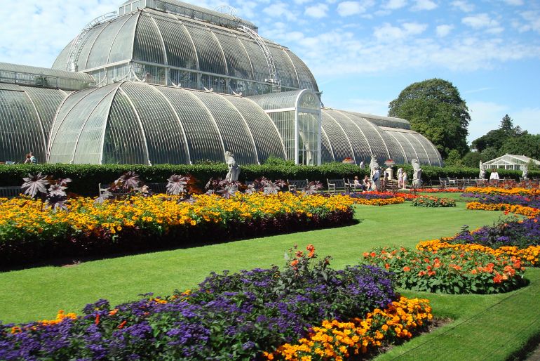 Lush garden with vibrant flowers in front of a large glass greenhouse under clear blue skies.
