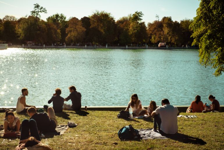 People relaxing by a sunlit park lake with sparkling water, surrounded by trees with autumn foliage.