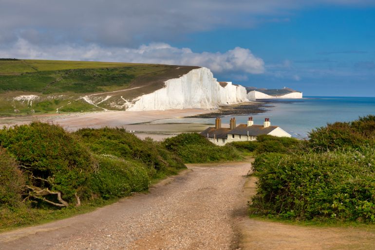 Mud path leading towards a coastal view with white chalk cliffs, blue ocean, greenery, and a house with red chimneys under a partly cloudy sky.