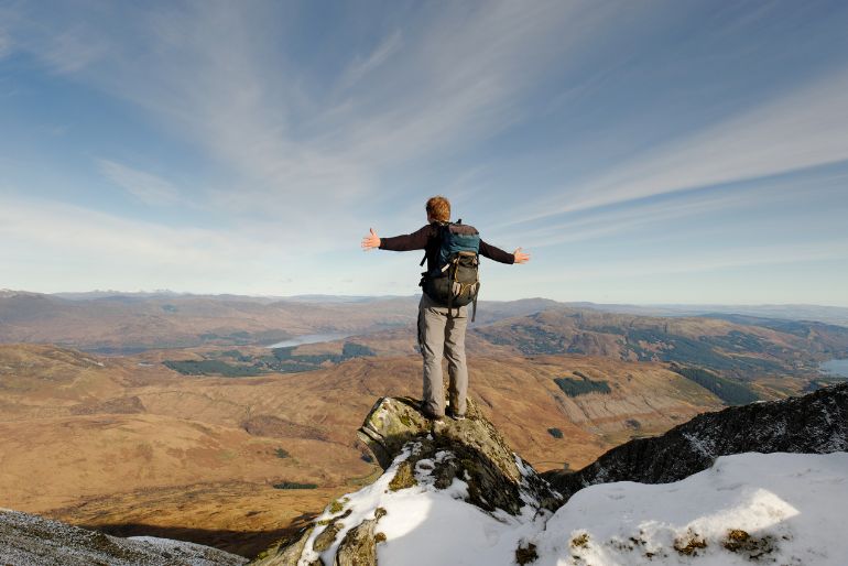 Hiker with arms outstretched stands on a mountain peak overlooking a vast valley with lakes and rolling hills, under a clear sky.