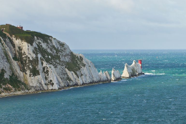White chalk cliffs with grassy tops beside a rough blue sea, under a partly cloudy sky, with a red navigation buoy offshore.