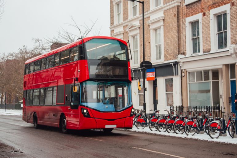 Red double-decker bus on snowy street next to bike-sharing station, with terraced houses in background.