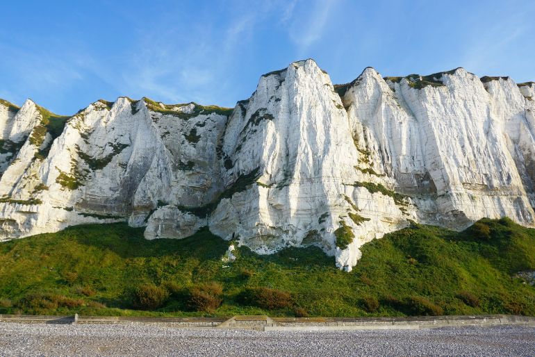 Imposing white chalk cliffs rise starkly against a blue sky above a pebble beach and a band of green vegetation.